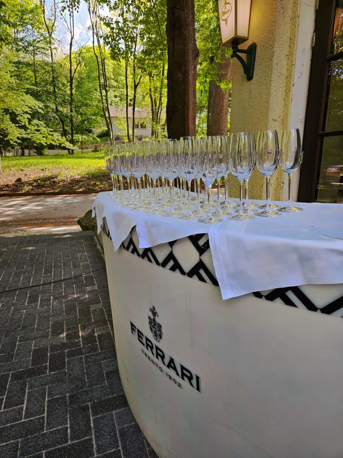Reihen von Sektgläsern auf einem eingedeckten Stehtisch vor dem Haus, dahinter der Wald