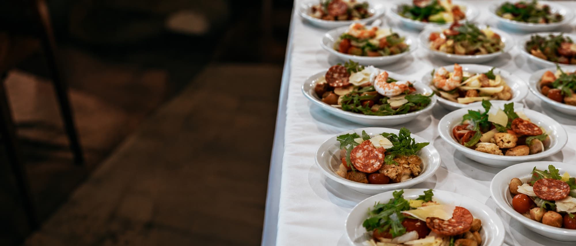 Reihen kleiner Vorspeisenschalen mit Salat, Salami und Garnelen auf einer weiß eingedeckten Buffettafel