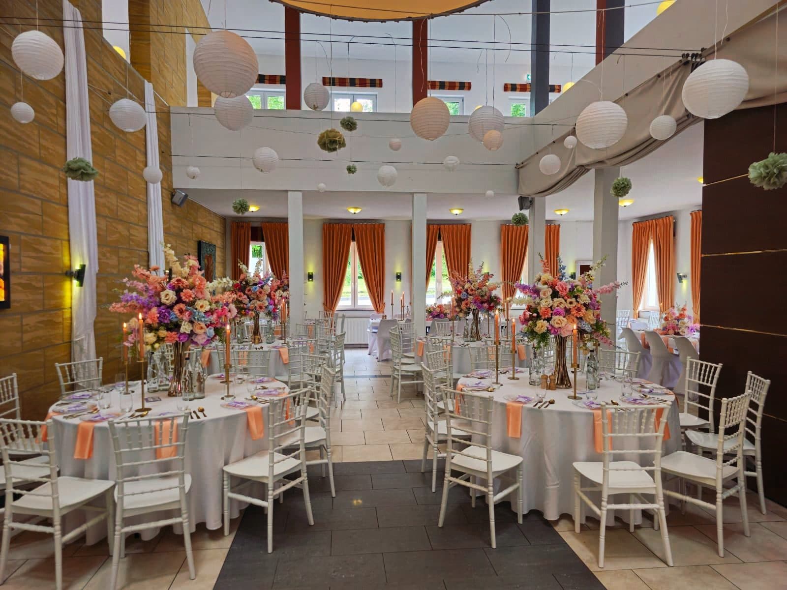 Festlich gedeckter Saal mit runden Tafeln, weißen Stühlen und hohem Blumenschmuck